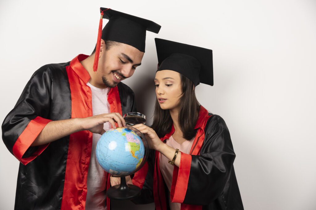 Pair of happy students in gown looking at globe on white background